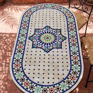 May include: Oval mosaic tile table with a detailed geometric pattern in blue, green, red, and yellow. The table has a white background with a dark brown metal frame and is surrounded by red brick flooring.