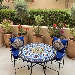 May include: A round mosaic tile table with a black metal frame and two matching chairs with blue cushions. The table has a detailed blue, white, yellow, and green pattern. The chairs have decorative pillows. Potted plants and flowers are in the background.
