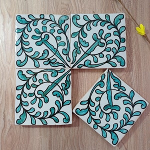 Könnte beinhalten: Eine Sammlung weißer Keramikfliesen mit einem türkis-schwarzen floralen Design. Die Fliesen sind quadratisch und rautenförmig, angeordnet auf einer Holzoberfläche. Das türkisfarbene Design zeigt stilisierte Blätter und Ranken.