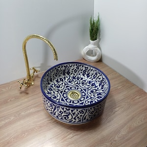 Może przedstawiać: Okrągła ceramiczna umywalka z niebiesko-białym kwiatowym wzorem. Umywalka ma mosiężny odpływ i jest połączona ze złotą baterią. W pobliżu znajduje się mały biały wazon z zielenią. To dekoracyjny element łazienki.