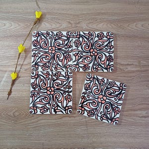 Op de afbeelding: Een set keramische tegels met een bloemenpatroon. De tegels zijn wit met een patroon van roze bloemen en zwarte contouren. Ze zijn gerangschikt op een houten oppervlak, met een takje gele bloemen op de achtergrond.