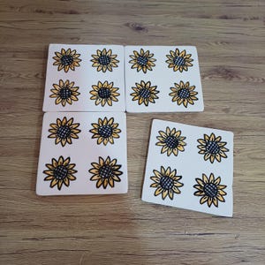Può includere: Un set di tre sottobicchieri quadrati bianchi, ognuno con un'illustrazione disegnata a mano di quattro girasoli gialli con centri scuri. I girasoli sono disposti in una griglia di due per due sulla superficie di ogni singolo sottobicchiere.
