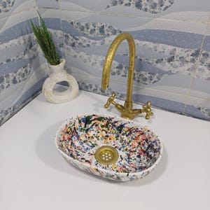 Puede incluir: Lavabo ovalado de cerámica con un diseño de pintura salpicada de colores. El lavabo se combina con un grifo dorado y un pequeño jarrón blanco con vegetación. El lavabo mide aproximadamente 50 cm de largo.
