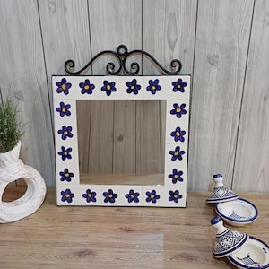 May include: A square wall mirror framed with white ceramic tiles featuring hand-painted blue flowers with yellow centers. The mirror is topped with a black wrought iron scrollwork accent piece. The frame consists of sixteen individual tiles.