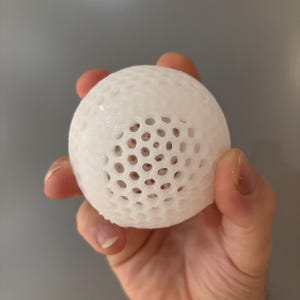 May include: A white, spherical object with a honeycomb-like pattern of holes. The object is held in a hand against a gray background. The ball appears to be made of a translucent material.