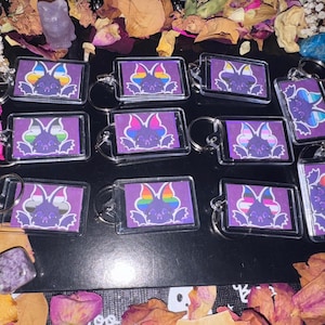 May include: A collection of rectangular keychains with a purple background and a bat design. The bat has rainbow-colored wings and is surrounded by a clear plastic frame. The keychains are on a dark surface with dried flowers and crystals.
