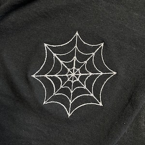 May include: Black fabric featuring a white embroidered spiderweb design. The spiderweb is centrally placed and has a detailed, symmetrical pattern. The fabric appears to be a soft material.