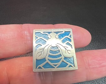 Puede incluir: Un colgante de plata con un diseño de abeja de esmalte azul. La abeja está delineada en plata y tiene un diseño detallado. El colgante tiene forma cuadrada y tiene un marco de plata.