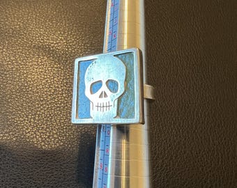 Puede incluir: Un anillo de plata con un diseño de calavera cuadrada de esmalte azul. El anillo está en un medidor de anillos.