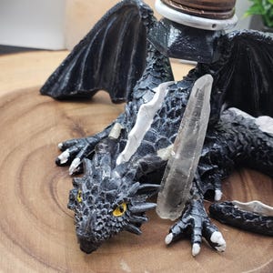 May include: A detailed black dragon figurine with yellow eyes and white accents, holding a clear crystal. The dragon is resting on a wooden surface. A decorative sphere and coasters are on the dragon's back.