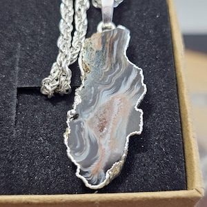 Op de afbeelding: Een zilveren ketting met hanger met een natuurlijke agaatsteen. De steen heeft een unieke, onregelmatige vorm met een zilverkleurige rand. De agaat toont een mix van grijze, witte en bruine kleuren, waardoor een marmer effect ontstaat. De ketting is ook zilver.