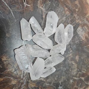 Puede incluir: Puntas de cristal de cuarzo transparente dispersas sobre una superficie de madera marrón oscuro y beige. Los cristales translúcidos tienen extremos puntiagudos y tamaños variables. La superficie de madera presenta un patrón de vetas natural.