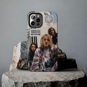 Puede incluir: Una funda negra para iPhone con una foto de la banda Nirvana. La foto muestra a los miembros de la banda Kurt Cobain, Krist Novoselic y Dave Grohl. La funda también tiene una pegatina de una cara sonriente con una X sobre los ojos.