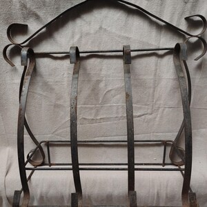 May include: A wrought iron magazine rack with a distressed finish. The rack features a curved top with decorative scrollwork and three vertical bars for holding magazines. The base has horizontal bars for additional support. The metal has a rustic, aged appearance.