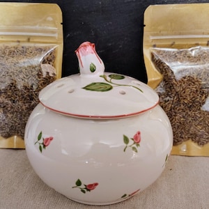 May include: A white ceramic jar with a rose-shaped lid and small pink rose accents. Two clear plastic bags filled with brown seeds are on either side of the jar. The jar is likely for storing spices or herbs.