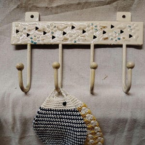 May include: Off-white metal wall hook with four hooks. The top bar has decorative cutouts and floral and blue accents. A small, crocheted item with navy and yellow details hangs from one hook.