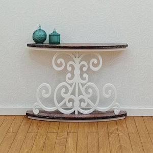 May include: A white, ornate console table with a dark brown top and base. Two teal glass jars sit on the table. The table has a decorative, scrolled design. The table is against a white wall and sits on a wooden floor.
