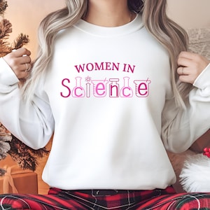 Pode incluir: Camisola branca com o texto "Women in Science" em letras cor-de-rosa. A palavra "Science" é composta por béqueres e tubos de ensaio estilizados.