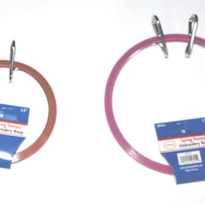 May include: Two circular spring tension embroidery hoops with silver metal tension springs. The smaller hoop is copper-colored, and the larger hoop is magenta. Each hoop features a blue product tag labeled Spring Tension Embroidery Hoop.