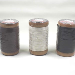 May include: Three spools of thread in varying colors. The spools have wooden tops and bottoms with a label. The thread colors are dark brown, white, and black. The thread is wrapped in clear plastic.