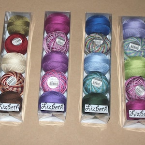 May include: Six clear plastic boxes each containing five spools of colorful embroidery thread. The spools are in shades of pink, purple, blue, green, and yellow. Each box has a label that reads "Lizbeth".