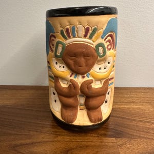 May include: A cylindrical ceramic vase with a black rim and a tan body. The vase features a relief design of a face with outstretched arms, and colorful accents in blue, yellow, and red. The vase is on a wooden surface.