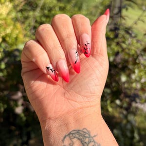 May include: A hand with long, almond-shaped nails. The nails feature a gradient design, transitioning from a nude base to a vibrant red at the tips. Delicate black leaf designs adorn each nail, adding a touch of elegance. A tattoo is visible on the wrist.
