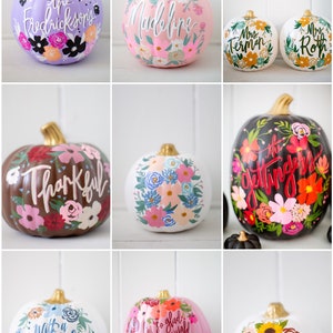 May include: A collection of nine decorative pumpkins featuring hand-painted floral designs and calligraphy. The pumpkins are painted in various colors including lavender, pink, white, brown, and black, each topped with a metallic gold-colored stem.