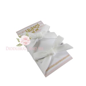 May include: Two white velvet hair bows on a pink and white striped card with the text "Diddlebugs and Me" and a website address.
