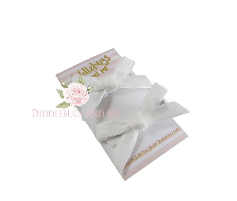 May include: Two white velvet hair bows on a pink and white striped card with the text "Diddlebugs and Me" and a website address.