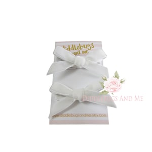 May include: Three white velvet hair bows with alligator clips. The bows are arranged on a white card with the text "Diddlebugs and Me" and a website address.