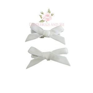May include: Two white satin ribbon bows with clips attached. The bows are tied in a simple knot and are perfect for adding a touch of elegance to any outfit.