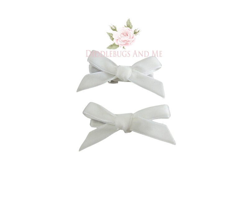 May include: Two white satin ribbon bows with clips attached. The bows are tied in a simple knot and are perfect for adding a touch of elegance to any outfit.