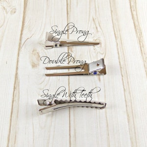 May include: Three silver hair clips on a wooden surface. The top clip is labeled "Single Prong", the middle clip is labeled "Double Prong", and the bottom clip is labeled "Single With Teeth".