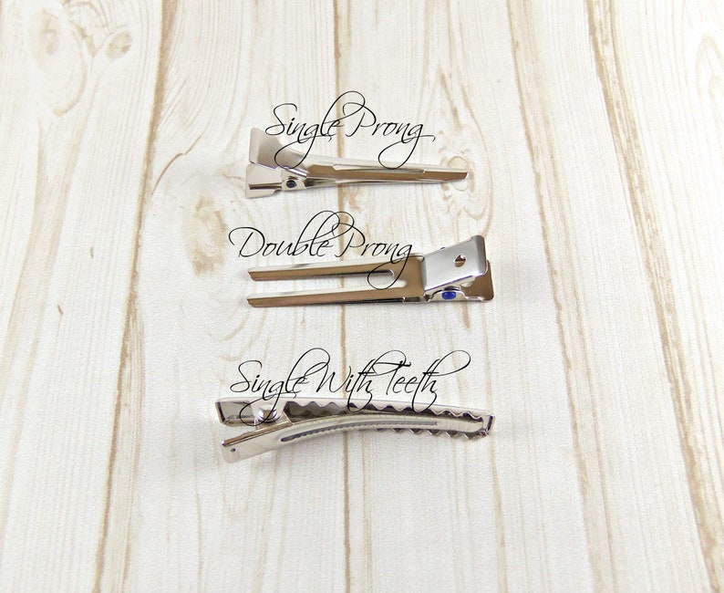 May include: Three silver hair clips on a wooden surface. The top clip is labeled "Single Prong", the middle clip is labeled "Double Prong", and the bottom clip is labeled "Single With Teeth".