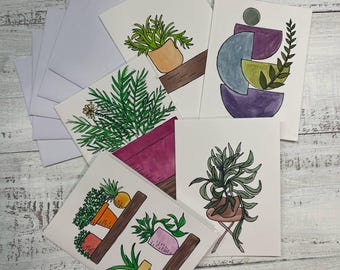 May include: A collection of watercolour art cards depicting potted plants and abstract designs. The cards show various green plants in orange, pink, and brown pots. A stack of white envelopes is visible on the left. The cards are on a light wood surface.
