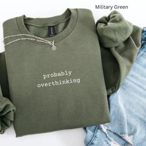 May include: A military green crewneck sweatshirt featuring the white text 'probably overthinking' embroidered on the chest. A silver-toned chain necklace with a small circular pendant rests on the neckline of the garment.