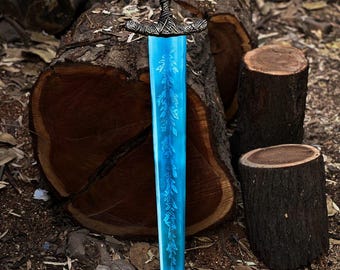 May include: A fantasy sword with a bright blue blade and ornate silver hilt. The blade features a detailed leaf pattern. The handle is wrapped in a dark, textured material. The sword is displayed against a backdrop of wooden logs and forest floor.