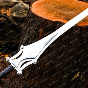May include: A silver sword with a broad blade and decorative hilt. The hilt has a detailed design with rounded features and a black leather-wrapped grip. The sword is displayed on a wooden surface.