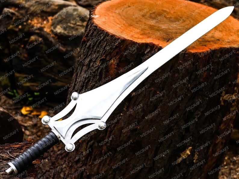 May include: A silver sword with a broad blade and decorative hilt. The hilt has a detailed design with rounded features and a black leather-wrapped grip. The sword is displayed on a wooden surface.