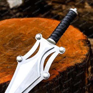 May include: A decorative sword with a silver blade and guard, and a black leather-wrapped handle. The sword is displayed on a wooden surface. The guard has four silver studs. The blade is approximately 30 cm long.