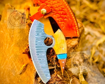 May include: A handcrafted cleaver knife with a patterned steel blade and a wooden handle with turquoise accents. The knife is paired with an orange leather sheath. The blade has a unique shape with a finger hole.