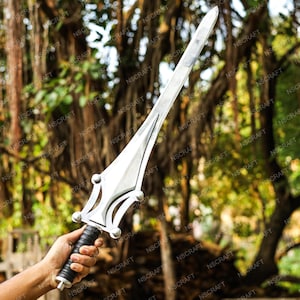 May include: A silver sword with a pointed blade and a decorative hilt. The hilt has a black grip and a stylized design. The sword is held against a blurred background of green foliage.