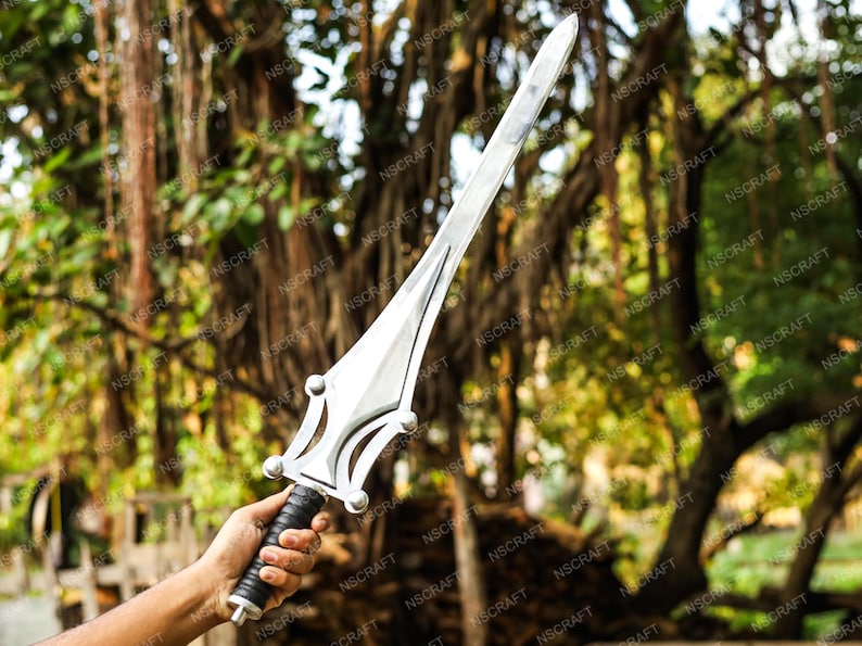 May include: A silver sword with a pointed blade and a decorative hilt. The hilt has a black grip and a stylized design. The sword is held against a blurred background of green foliage.