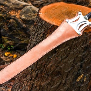 May include: A sword with a leather sheath rests on a tree stump. The sword has a silver crossguard and a black handle. The sheath is light brown and stitched along the edges. The sword is approximately 76 cm long.