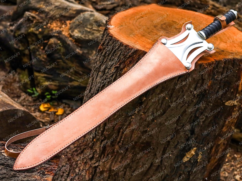 May include: A sword with a leather sheath rests on a tree stump. The sword has a silver crossguard and a black handle. The sheath is light brown and stitched along the edges. The sword is approximately 76 cm long.