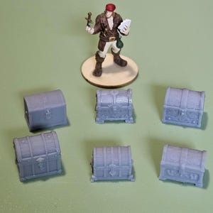 Może przedstawiać: Miniaturowa figurka osoby w brązowym płaszczu i czerwonym kapeluszu, trzymającej miecz i papier, stoi na drewnianej podstawie. Sześć małych, szarych skrzyń ze skarbami jest ułożonych wokół figurki na zielonej powierzchni. Skrzynie mają dekoracyjne detale.