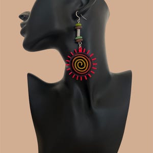May include: A pair of long, dangling earrings with a red, orange, and yellow sun design. The sun has a spiral in the center and is surrounded by red rays. The earrings also have a silver hook and a brown, green, and silver bead dangle.