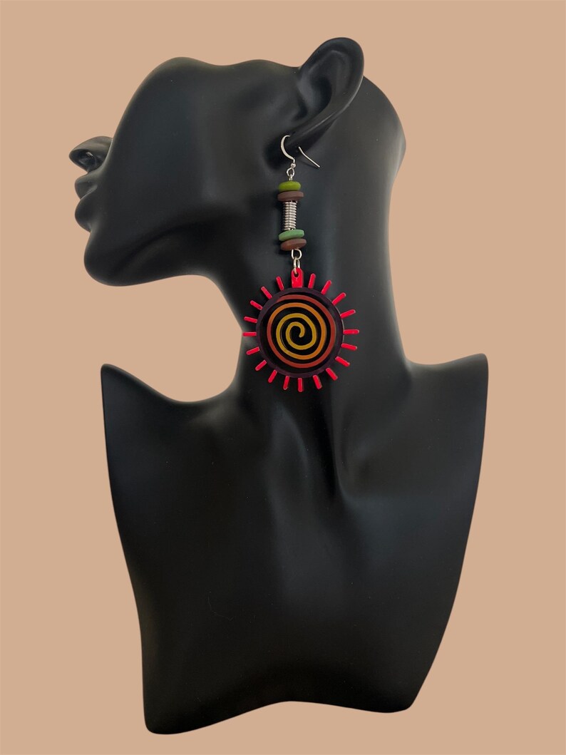May include: A pair of long, dangling earrings with a red, orange, and yellow sun design. The sun has a spiral in the center and is surrounded by red rays. The earrings also have a silver hook and a brown, green, and silver bead dangle.