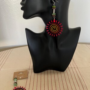 May include: A pair of handmade wooden earrings with a red, orange, and yellow sun design. The earrings are hanging from a silver hook and have a green, brown, and silver bead dangle.
